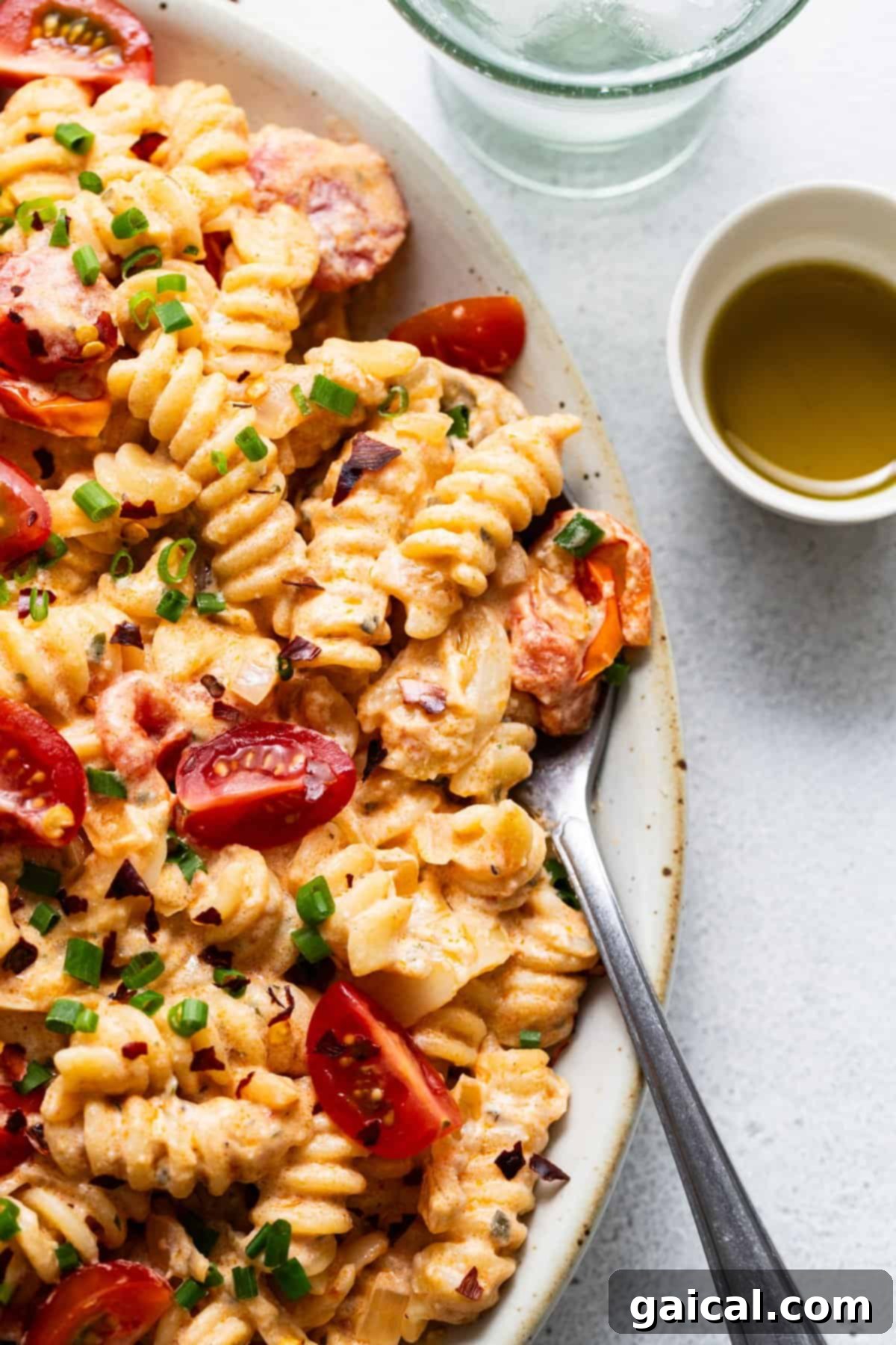 A dish of creamy pasta with sliced sausage, cherry tomatoes, and garnished with chopped green onions, served with a side of olive oil.