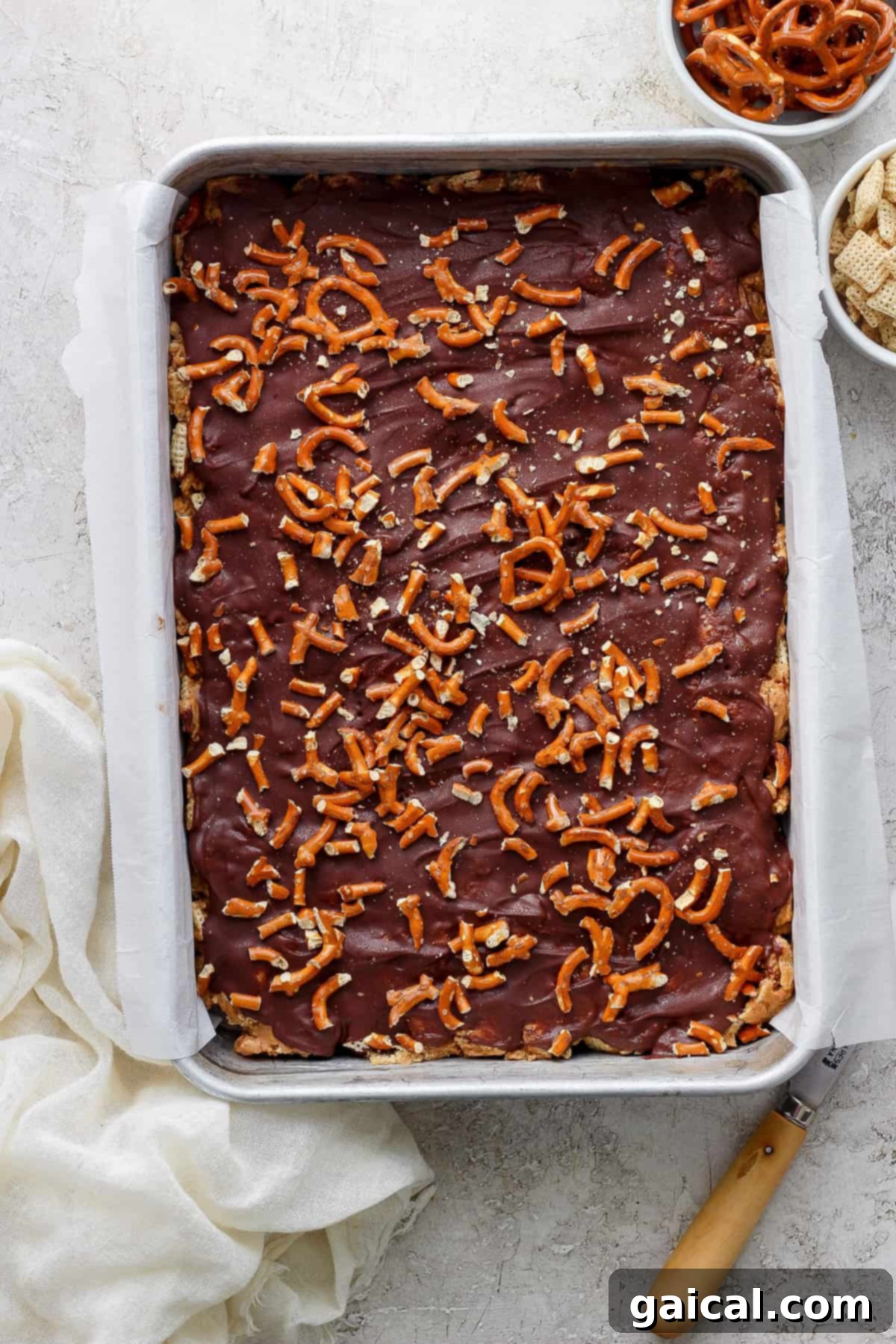 A tray of chocolate-covered toffee sprinkled with pretzel pieces on a kitchen counter, accompanied by a cloth and a spatula.