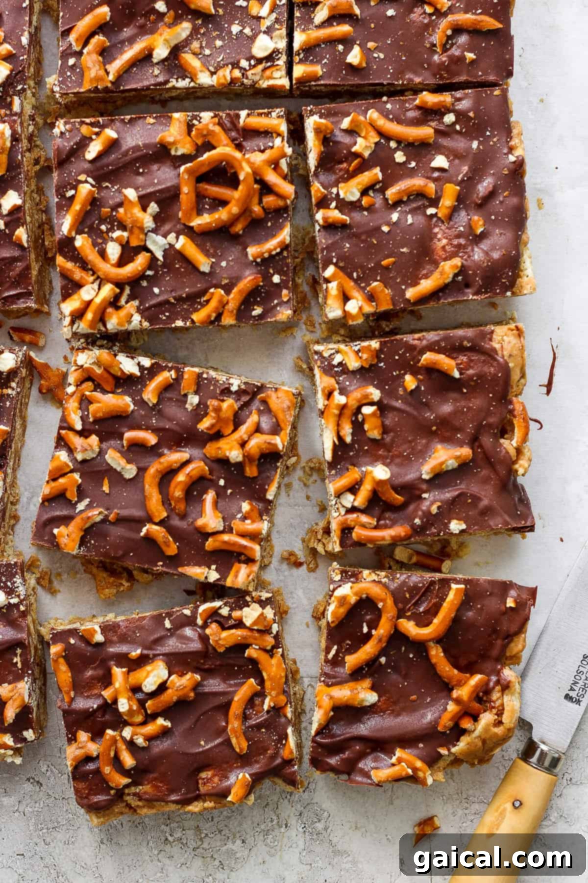 Chocolate-covered toffee bars topped with pretzel pieces on parchment paper, cut into rectangles, with a knife resting to the side.