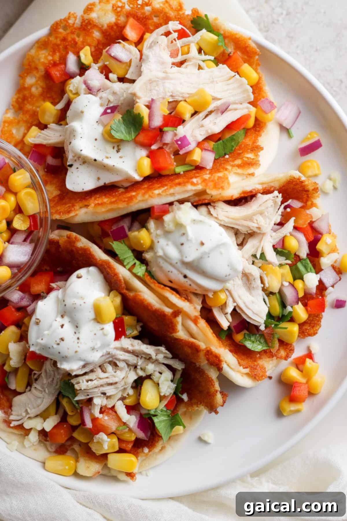 Zesty Cotija Chicken Tacos 2 A plate containing chicken tacos garnished with corn, diced tomatoes, red onion, cilantro, and a dollop of sour cream, served on a white surface.