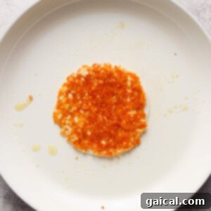 A single, fried hash brown resting on a white, circular non-stick pan with a red handle, displayed on a grey surface with a few scattered crumbs and oil droplets.