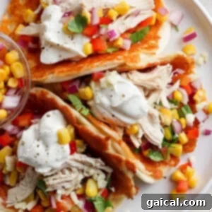 A plate containing chicken tacos garnished with corn, diced tomatoes, red onion, cilantro, and a dollop of sour cream, served on a white surface.