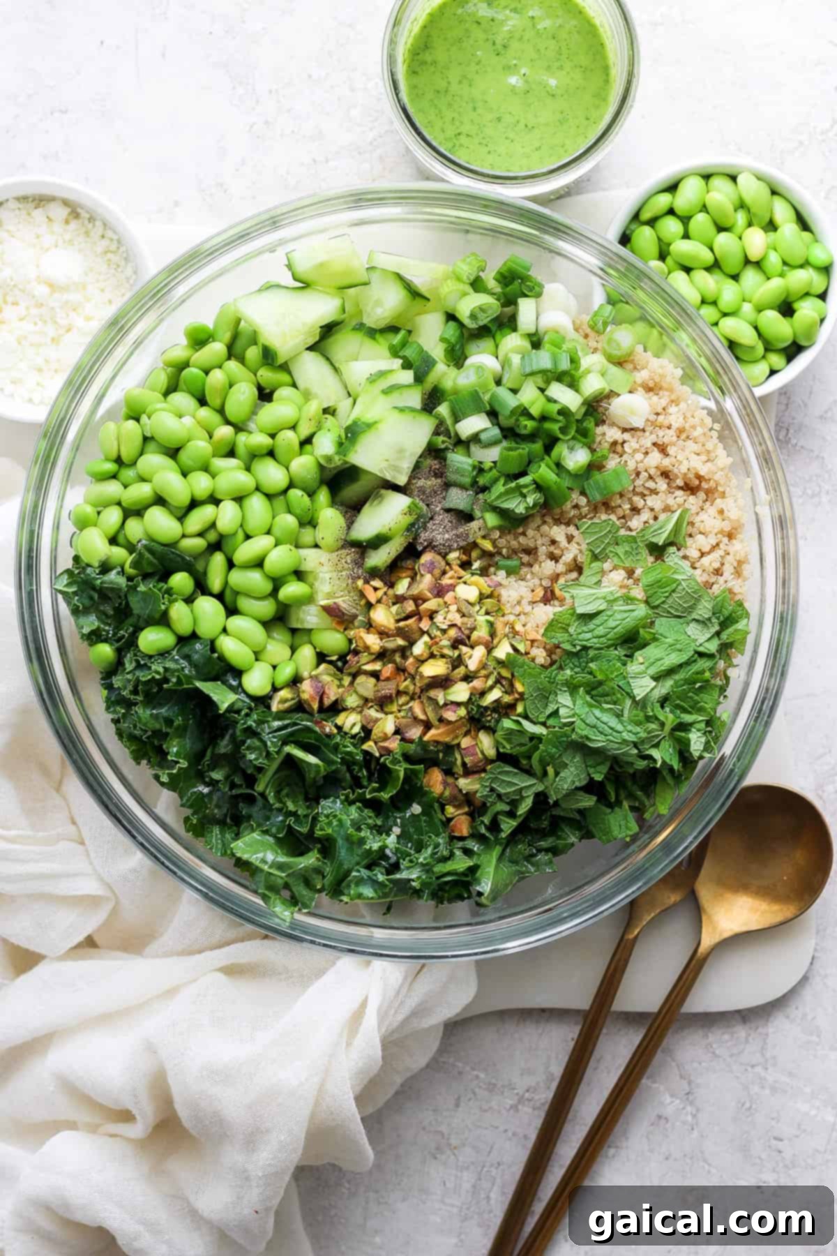 Vibrant Green Goddess Quinoa Bowl 2 A bowl filled with various green ingredients including kale, cucumbers, peas, and chopped nuts, with a sprinkle of grated cheese, accompanied by a jar of green dressing and a spoon on a white.