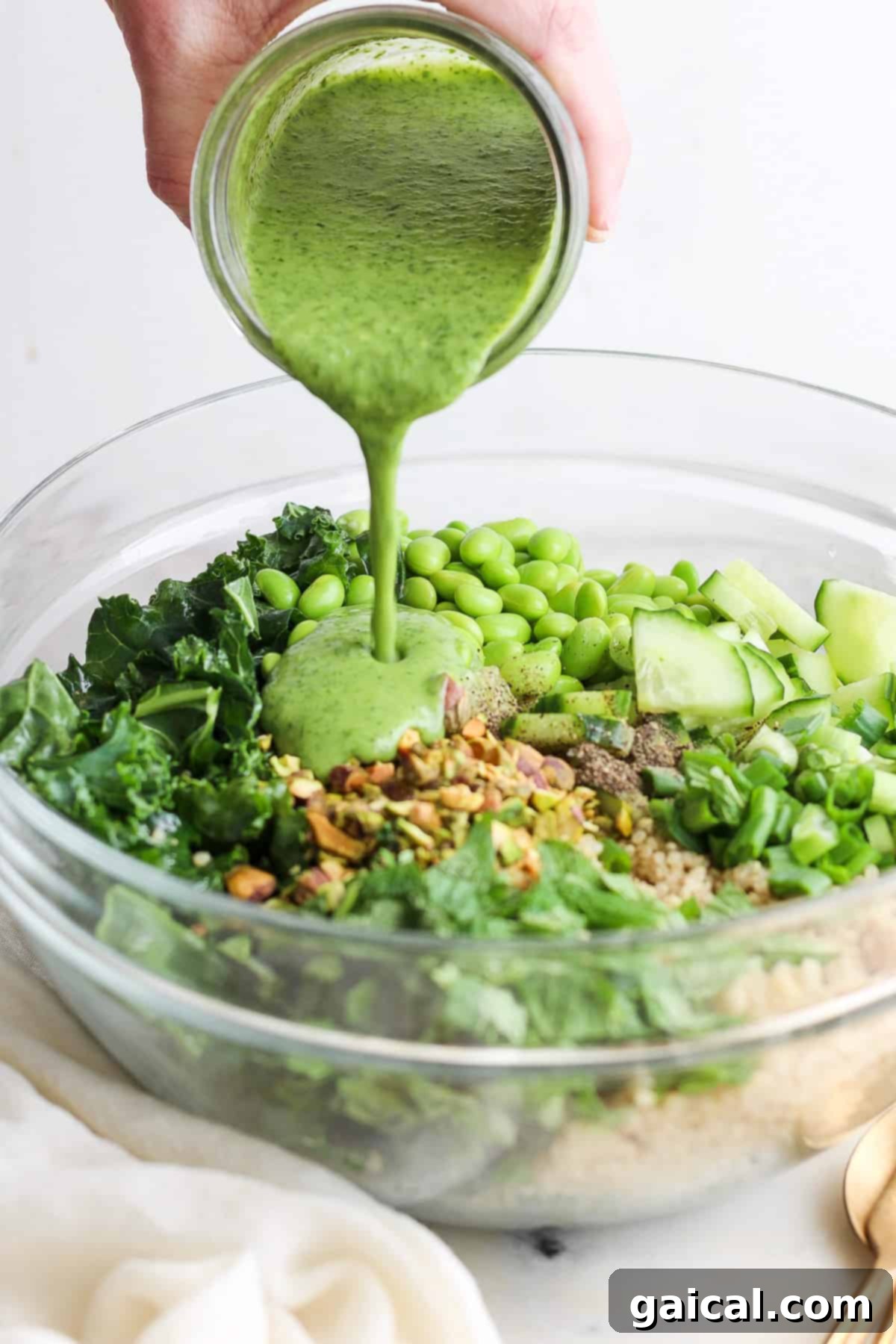 Vibrant Green Goddess Quinoa Bowl 6 Pouring a green dressing over a bowl of salad with kale, quinoa, cucumber, and green peas.
