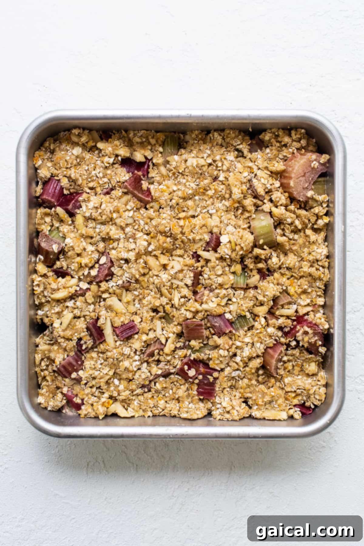 A metal baking pan filled with a half-assembled rhubarb crisp, showing the fruit filling topped with the oat crumble.