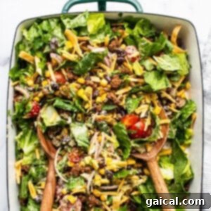 A colorful taco salad with lettuce, tomatoes, ground beef, cheese, and black beans served in a rectangular dish with wooden serving utensils.