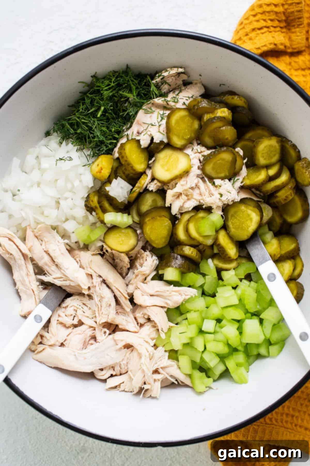 A vibrant and appealing display of fresh, key ingredients for the dill pickle chicken salad, presented in a large mixing bowl with tongs. Ingredients include perfectly shredded chicken, finely chopped white onions, bright green sliced baby dill pickles, and crisp diced celery, all awaiting combination.