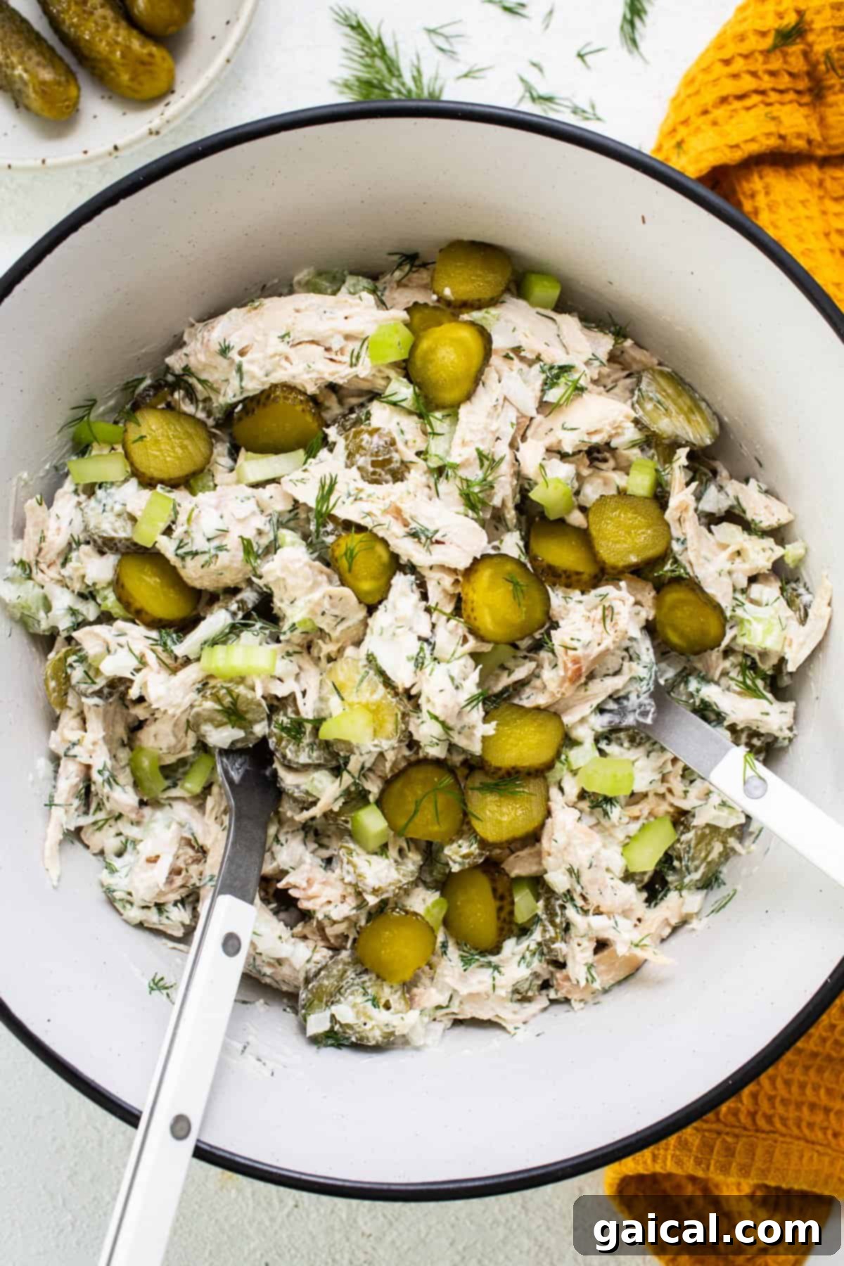 A beautifully composed bowl of creamy dill pickle chicken salad, artfully garnished with fresh dill sprigs and extra pickle slices, accompanied by elegant serving utensils, all resting on a rustic wooden surface, highlighting its appetizing readiness for enjoyment and showcasing its rich texture.