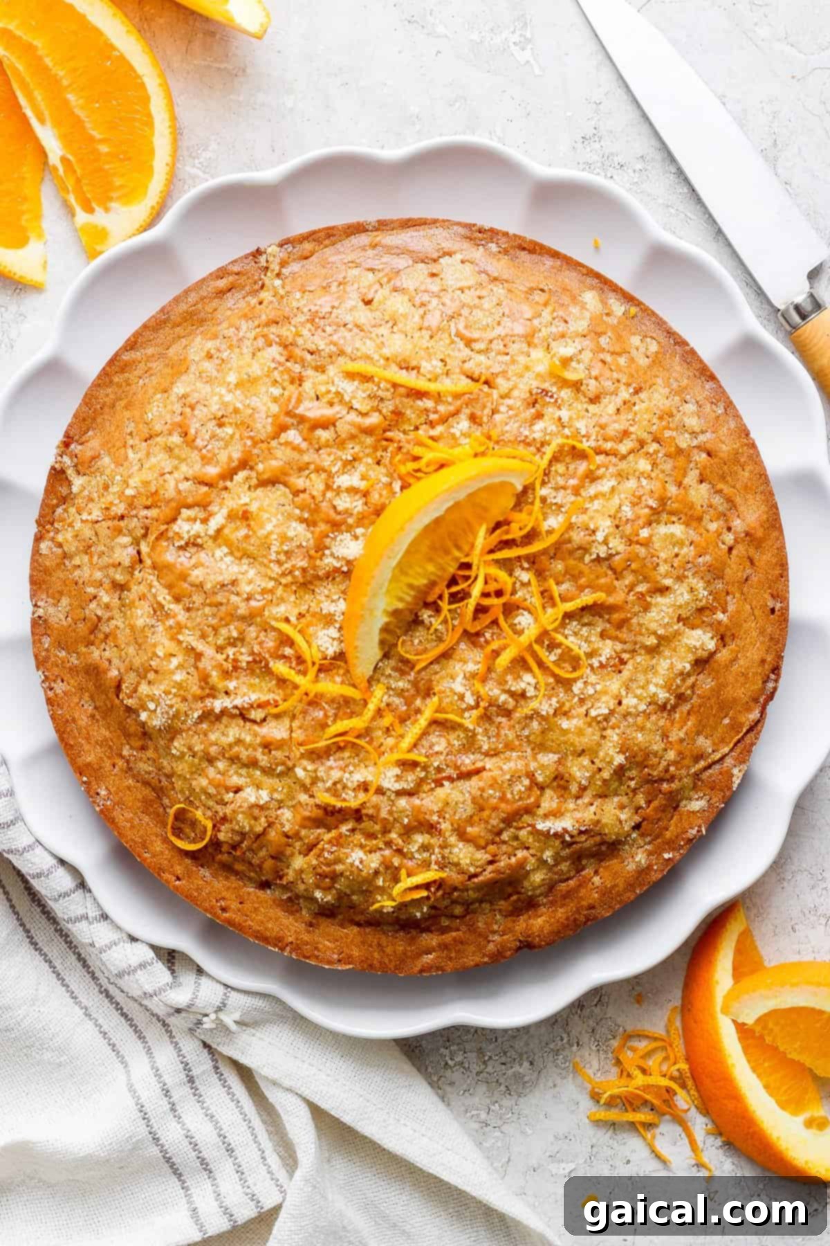 A beautifully baked orange olive oil cake, garnished with fresh orange zest, sits beside a scattering of fresh orange slices on a clean white plate, ready to be enjoyed.