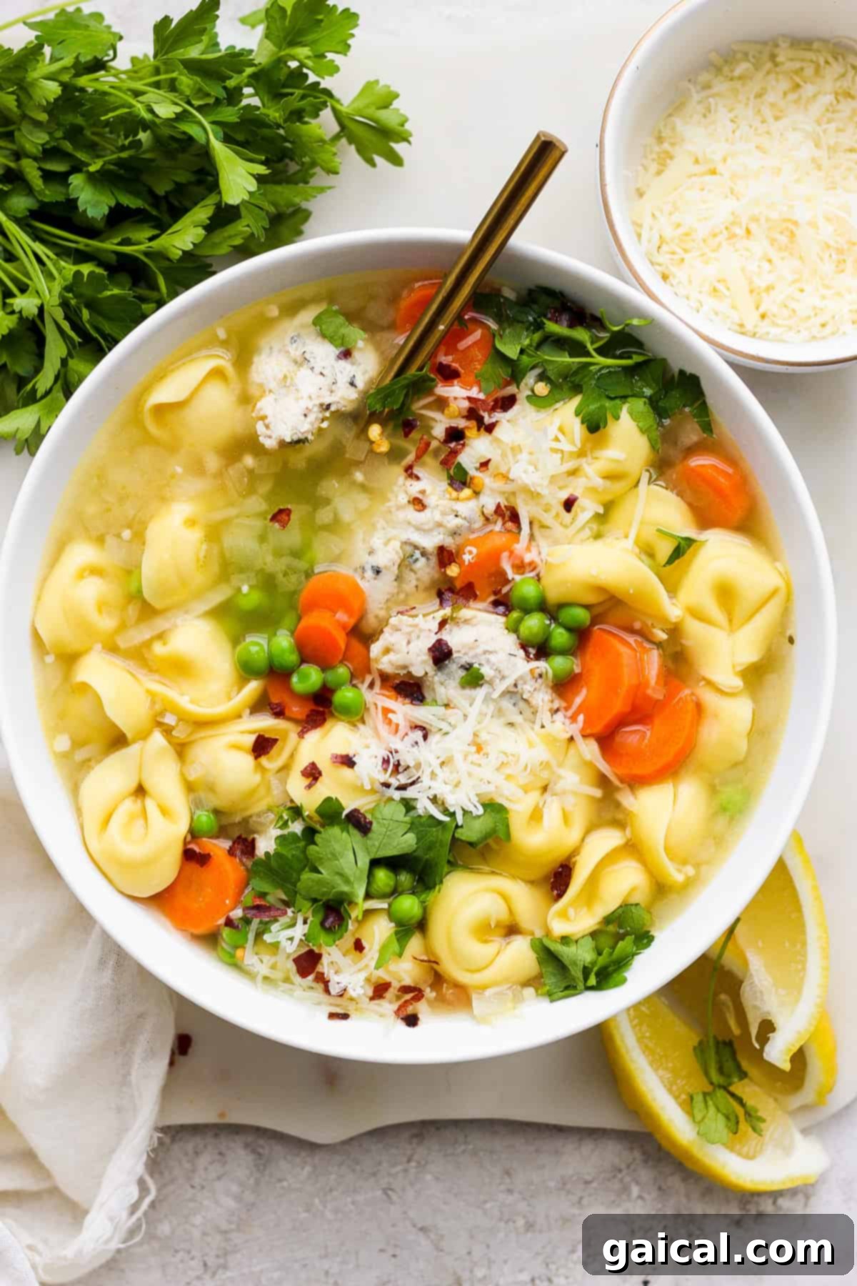 A bowl of chicken tortellini soup garnished with fresh parsley, shredded parmesan cheese, and a sprinkle of red pepper flakes, served with fresh lemon wedges on the side.