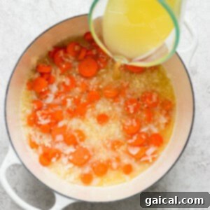 Pouring chicken broth into a pot with cooking onions and carrots.