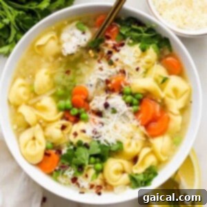 A bowl of tortellini soup with carrots, peas, and garnished with parsley and grated cheese.