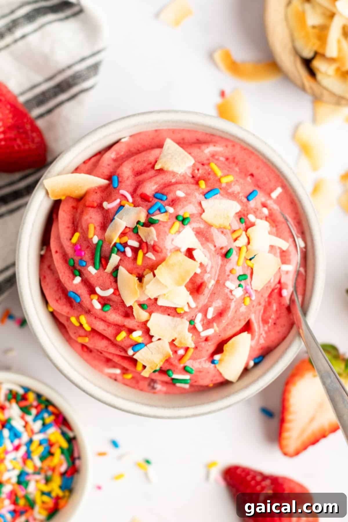 Strawberry Soft Swirl 2 A vibrant pink smoothie bowl topped with coconut flakes and rainbow sprinkles, accompanied by fresh strawberries and a spoon.