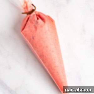 Strawberry Soft Swirl 10 A piping bag filled with pink frosting, tied at the top, resting on a white marble surface.