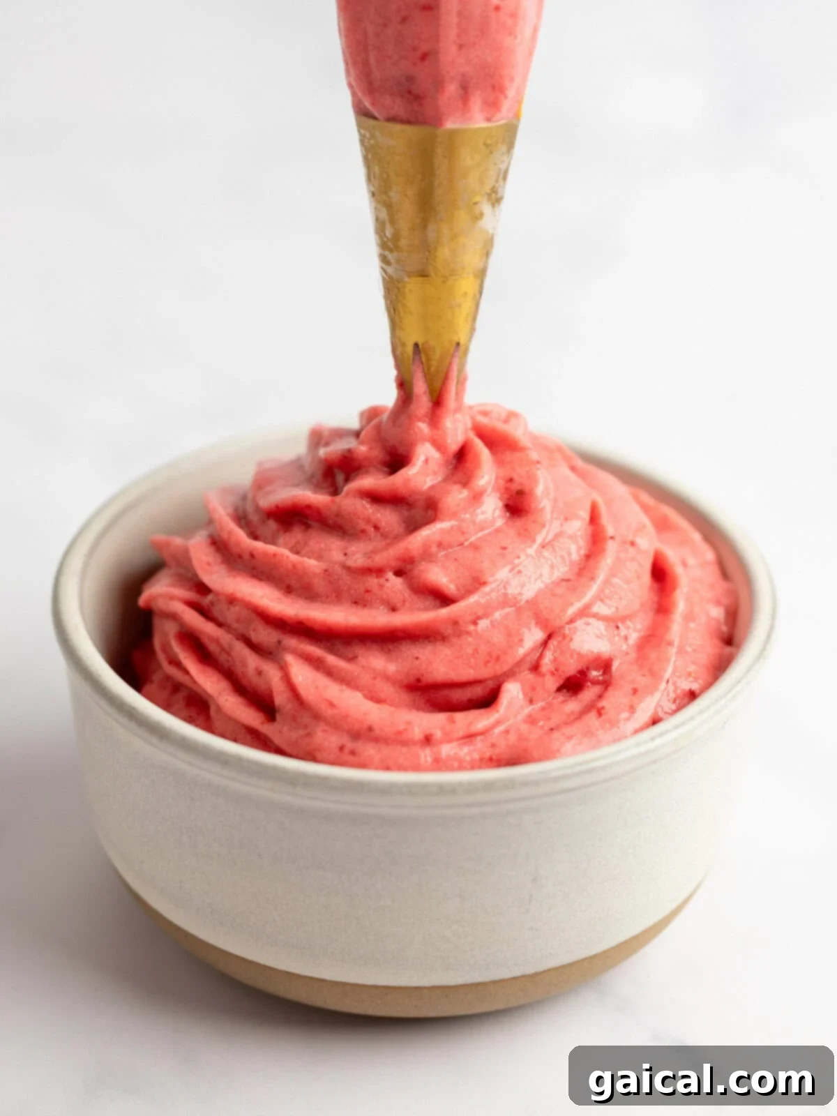 Strawberry Soft Swirl 5 A piping bag dispensing creamy strawberry frosting into a small beige bowl against a white background.
