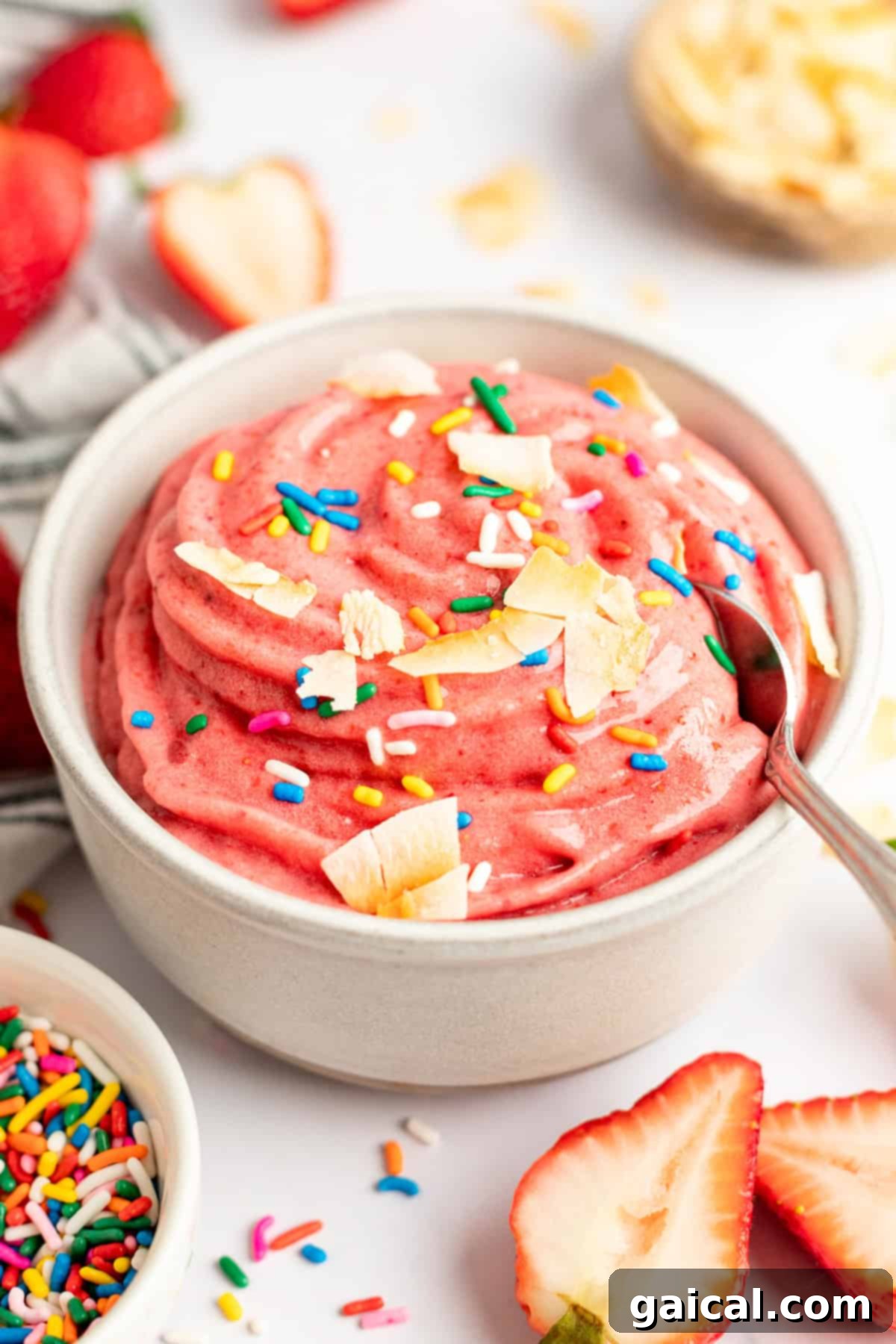 Strawberry Soft Swirl 6 A bowl of pink strawberry banana smoothie bowl topped with sprinkles and coconut flakes, surrounded by fresh strawberries and sprinkles.