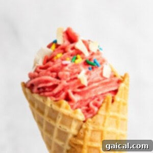 A close-up of a strawberry soft serve ice cream cone topped with sprinkles and white chocolate chunks.