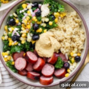A fully assembled Hodgepodge Protein Bowl, featuring layers of quinoa, colorful kale salad, sliced sausage, and a dollop of hummus, visually appealing and ready to eat.