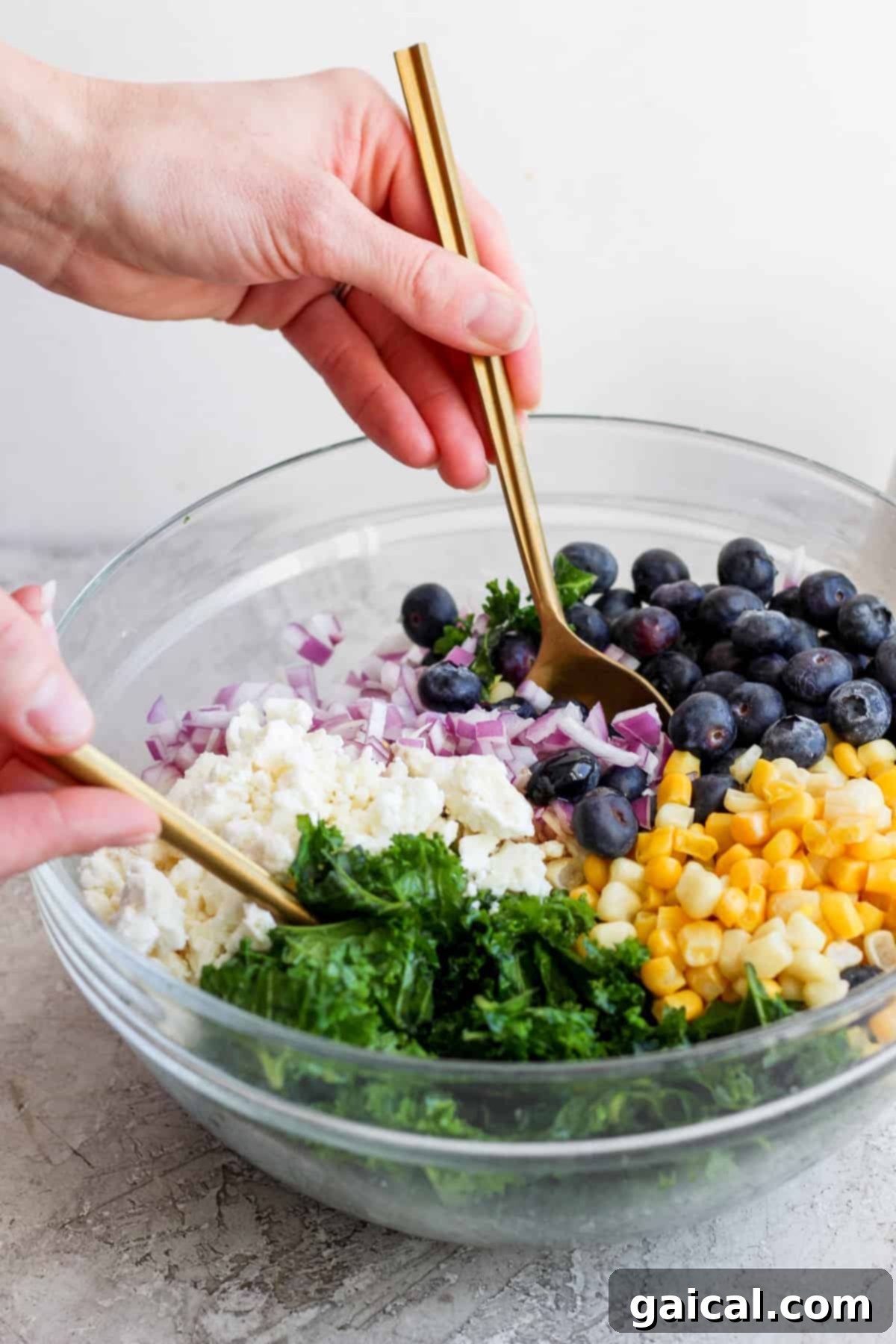 A person elegantly mixing a colorful and fresh salad, featuring bright blueberries, golden corn kernels, finely chopped red onions, and vibrant green kale, using golden tongs in a clear glass bowl.