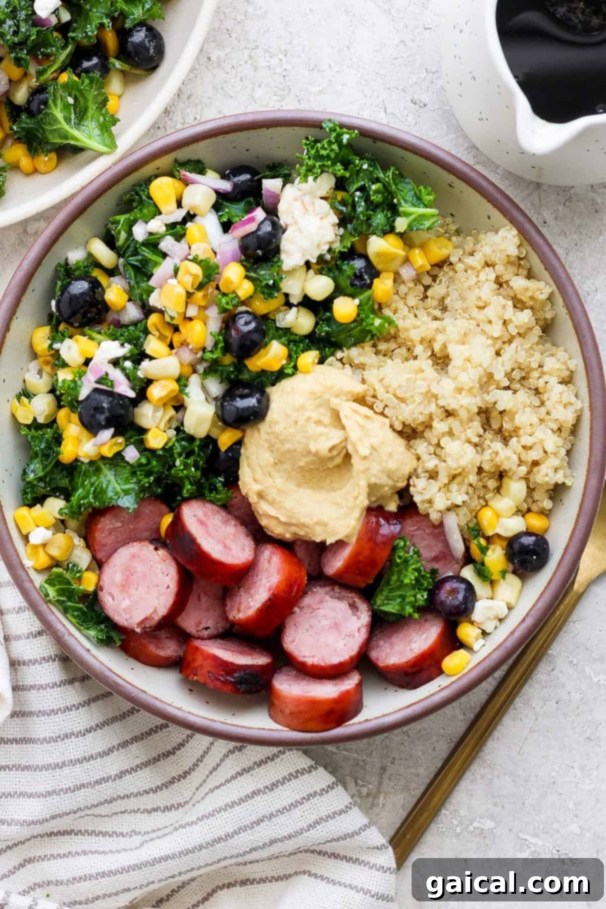 A meticulously arranged Hodgepodge Protein Bowl, showcasing fluffy quinoa, perfectly sliced sausage, a colorful mix of kale, corn, and blueberries, all crowned with a generous dollop of creamy hummus.