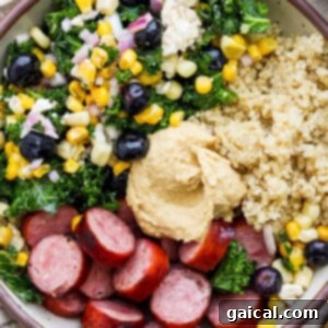A perfectly assembled bowl featuring quinoa, thinly sliced sausage, a rich mix of kale, corn, and black olives, beautifully topped with a smooth dollop of hummus.