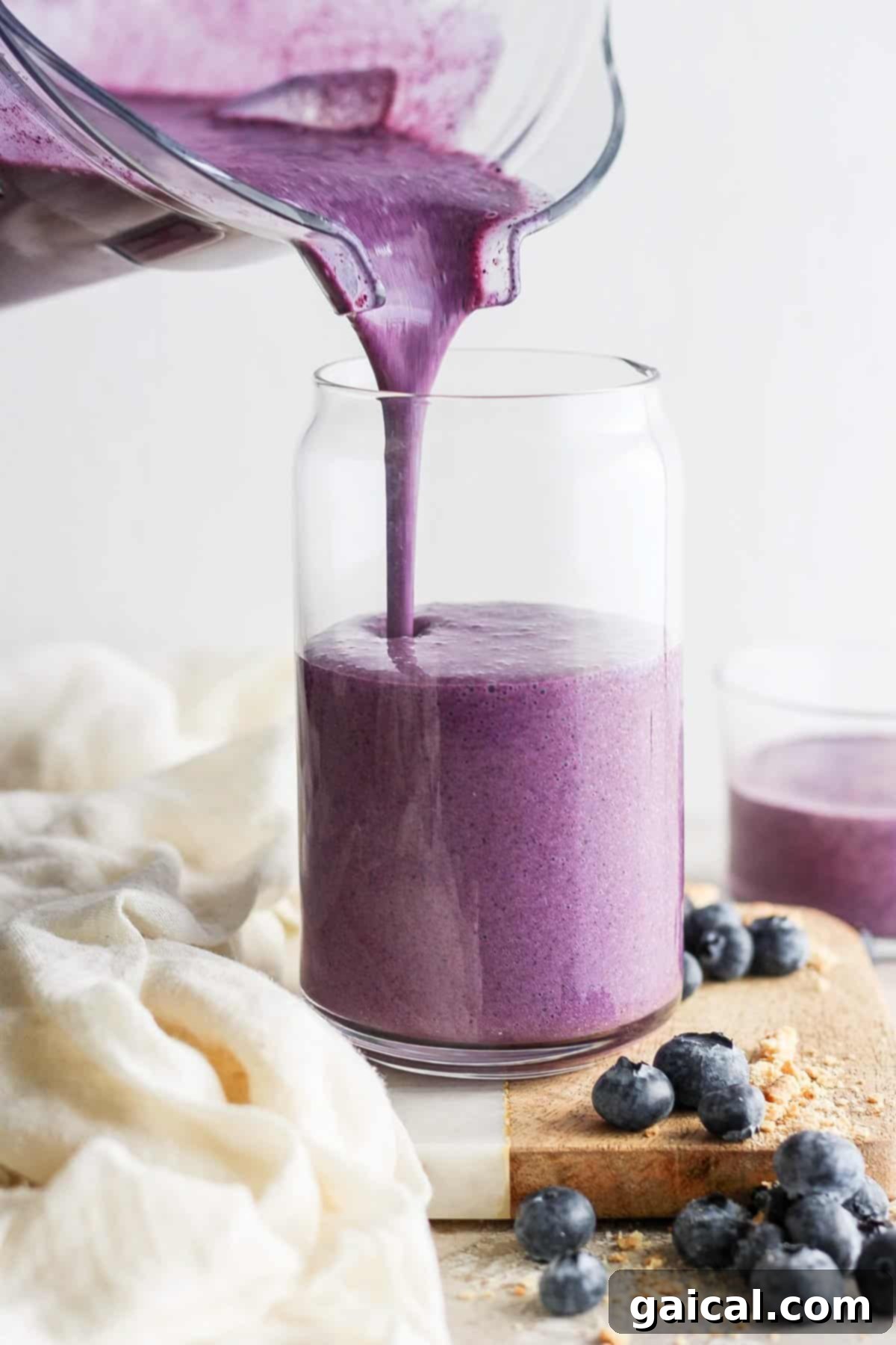 Vibrant Plate Collective 4 Pouring a fresh blueberry smoothie into a glass jar, surrounded by scattered blueberries on a wooden board.