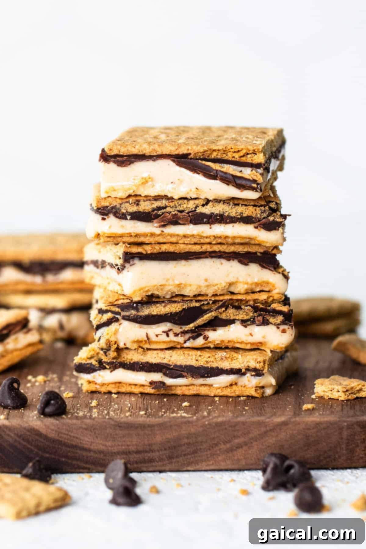Peanut Butter Protein Smores Freeze 7 A stack of golden s'mores bars with layers of graham crackers, creamy filling, and chocolate on a wooden board, with scattered chocolate chips around.