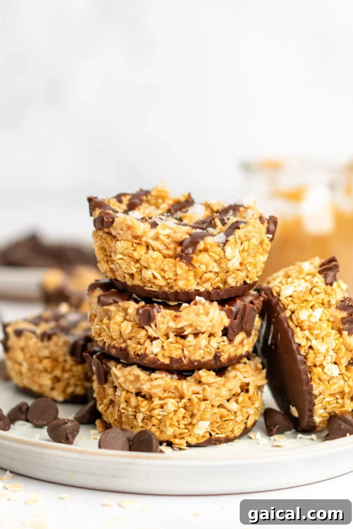 A stack of four oat and chocolate dessert bars on a plate with chocolate chips scattered around. A jar with peanut butter is visible in the background.