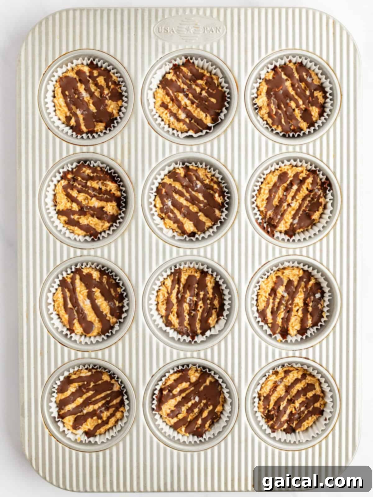 A muffin tin filled with 12 chocolate-drizzled oatmeal cookies in white paper liners.
