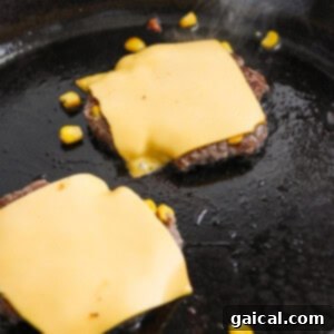 Confetti Corn Smash Burger 10 Two beef patties with melted cheese slices on top cooking in a skillet, showcasing the ideal doneness.