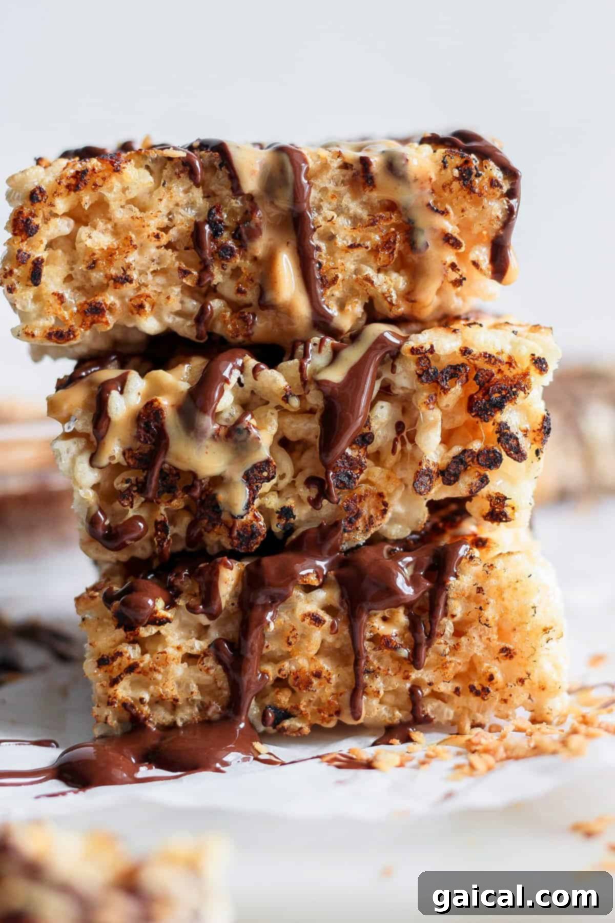 Stack of three toasted rice Krispie treats drizzled with chocolate and peanut butter on a white surface, garnished with chopped nuts.