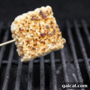 A square rice cake on a skewer is being grilled over open flames, showing light charring and crispness, reminiscent of toasted rice Krispie treats.