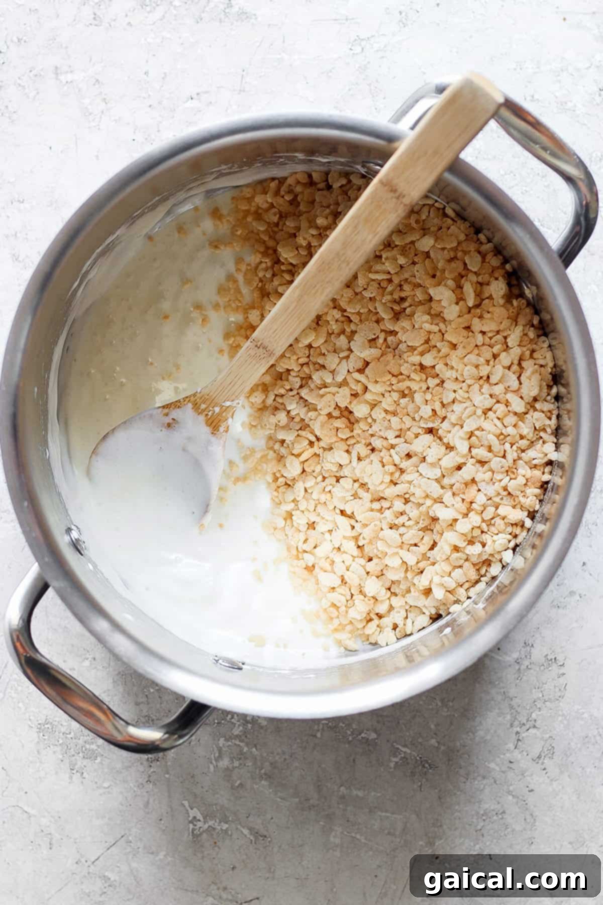A pot filled with melted marshmallows and some toasted Rice Krispie treats, with a wooden spoon resting inside, on a light-colored surface.