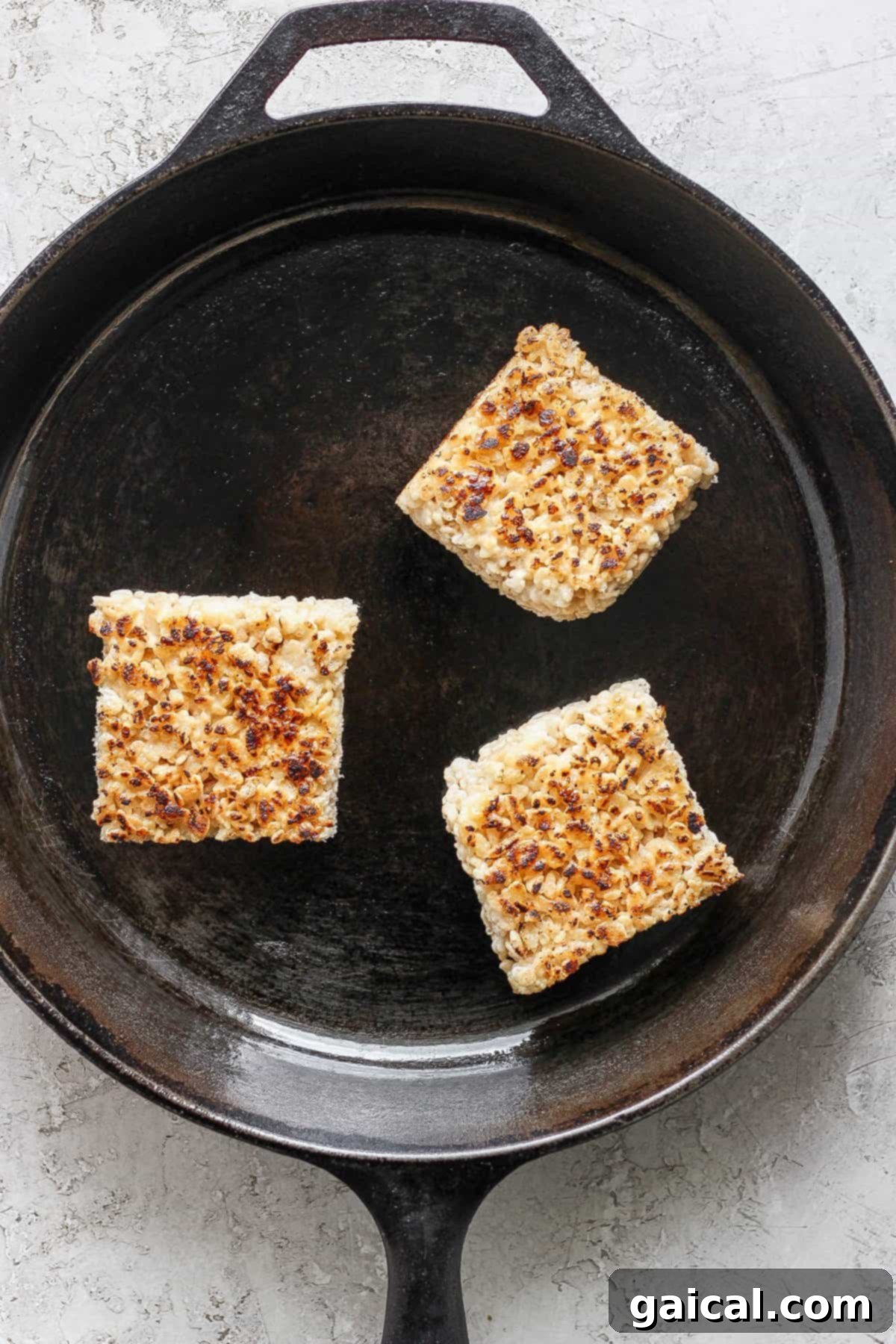 Three toasted Rice Krispie treats in a cast iron skillet on a light textured surface.