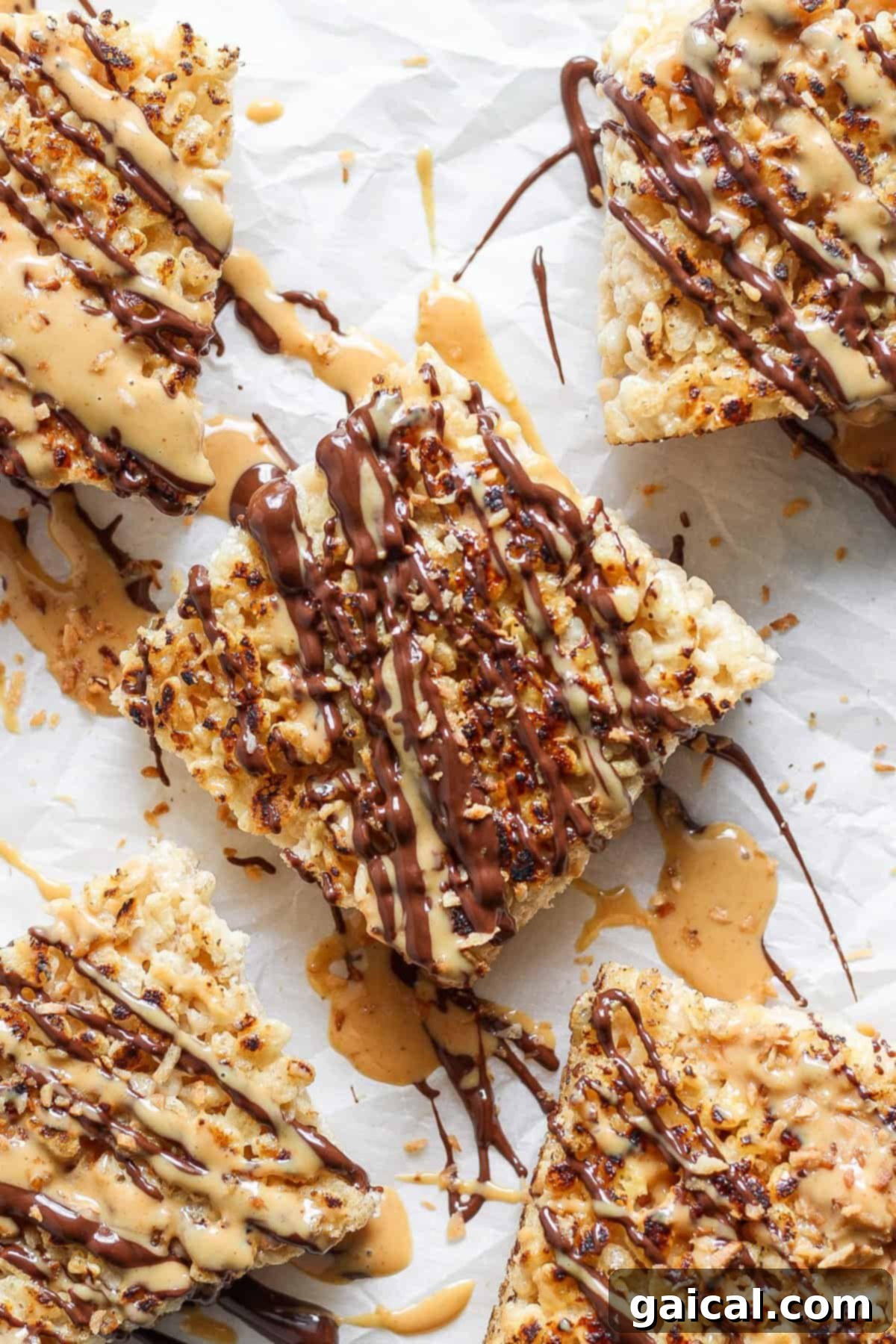 Close-up of five toasted Rice Krispie treats drizzled with chocolate and peanut butter, arranged on parchment paper. The treats are topped with crushed nuts.