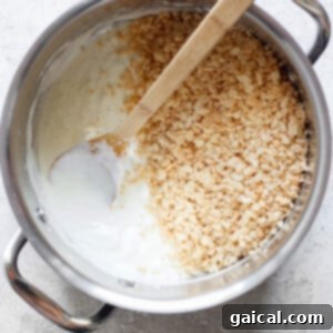 A pot filled with melted marshmallows and some toasted Rice Krispie treats, with a wooden spoon resting inside, on a light-colored surface.