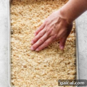 A hand pressing down on a tray filled with warm, toasted Rice Krispie treats mixture.