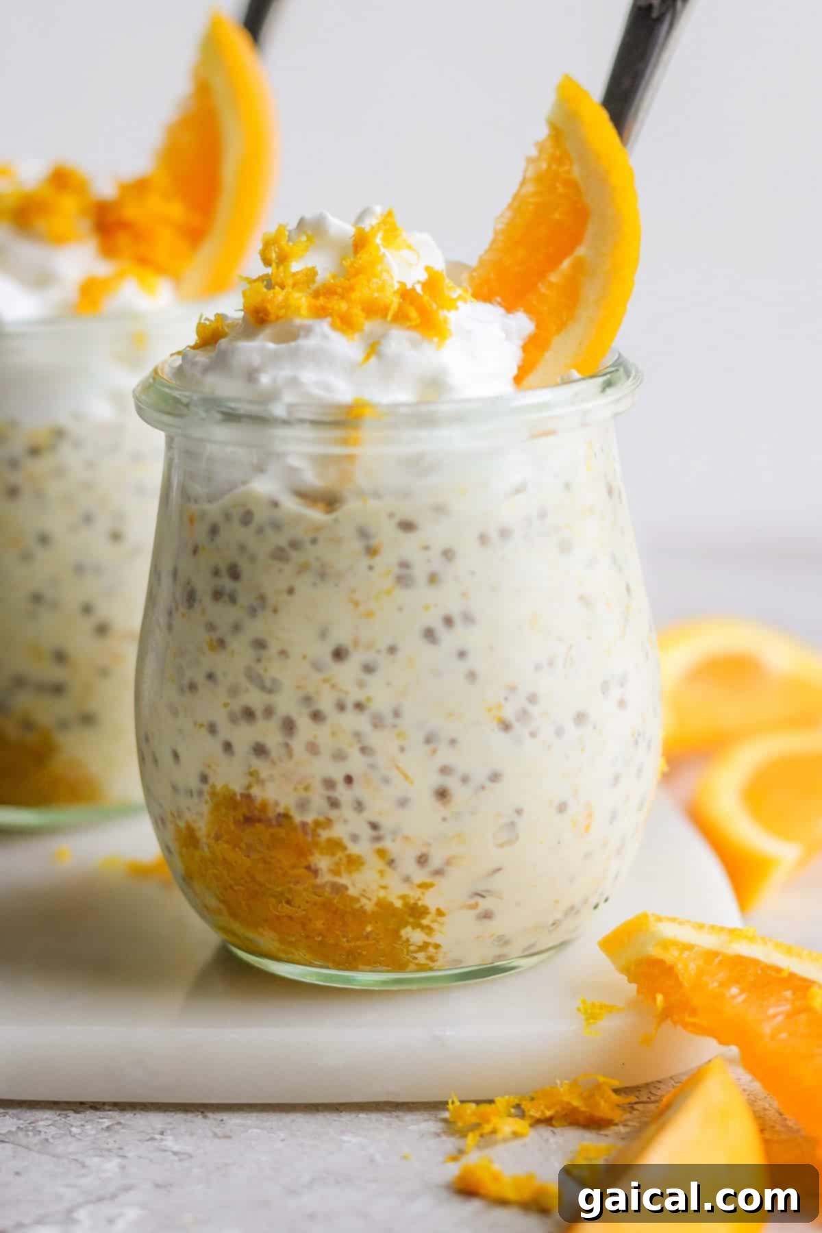 A glass jar filled with chia seed pudding topped with whipped cream, orange zest, and an orange slice, giving it a delightful creamsicle overnight oats feel, accompanied by pieces of orange on a white surface.