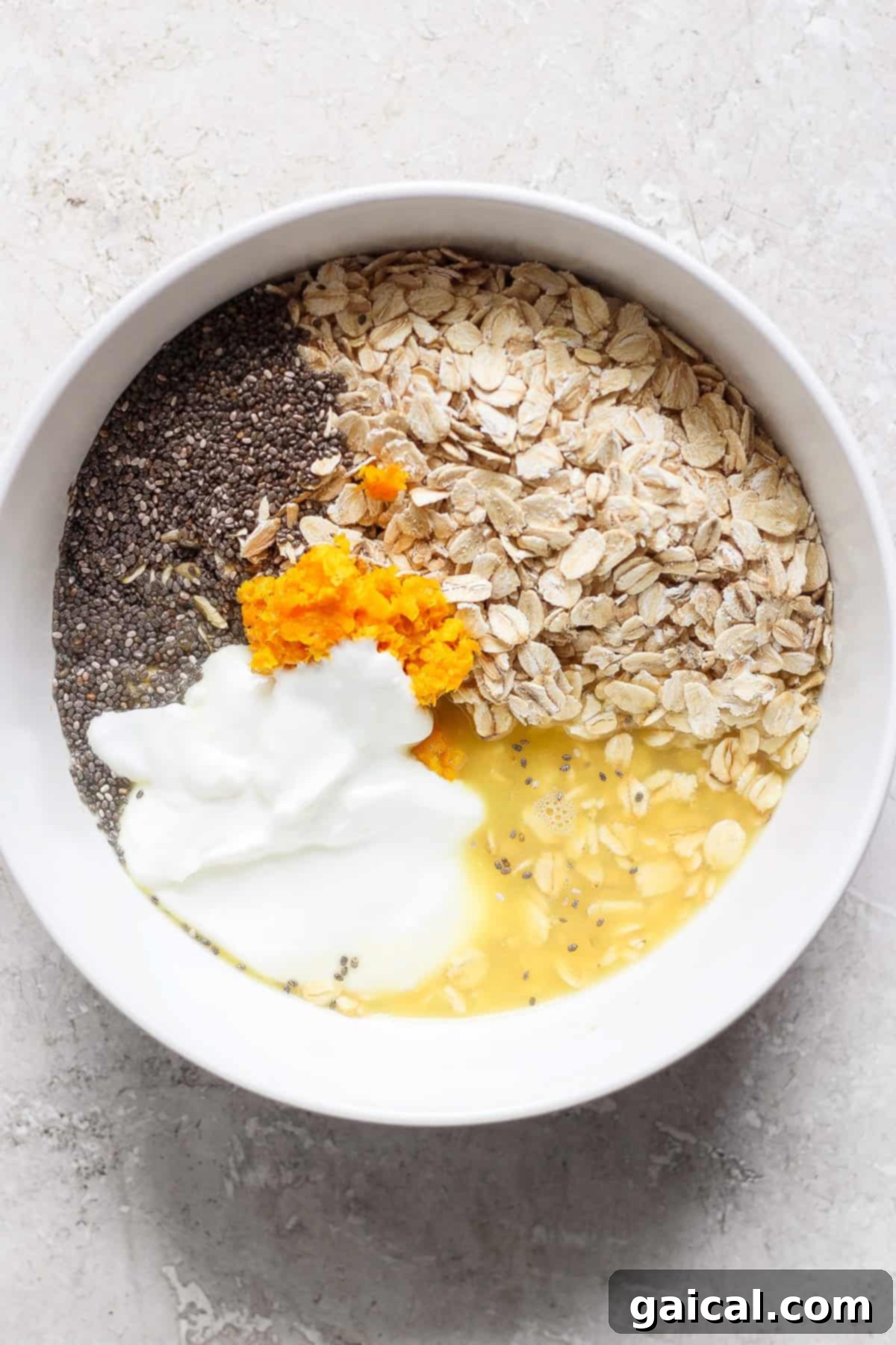 A white bowl contains oats, chia seeds, orange zest, yogurt, and liquid—likely milk or water—on a light-colored surface, creating a creamsicle overnight oats sensation.