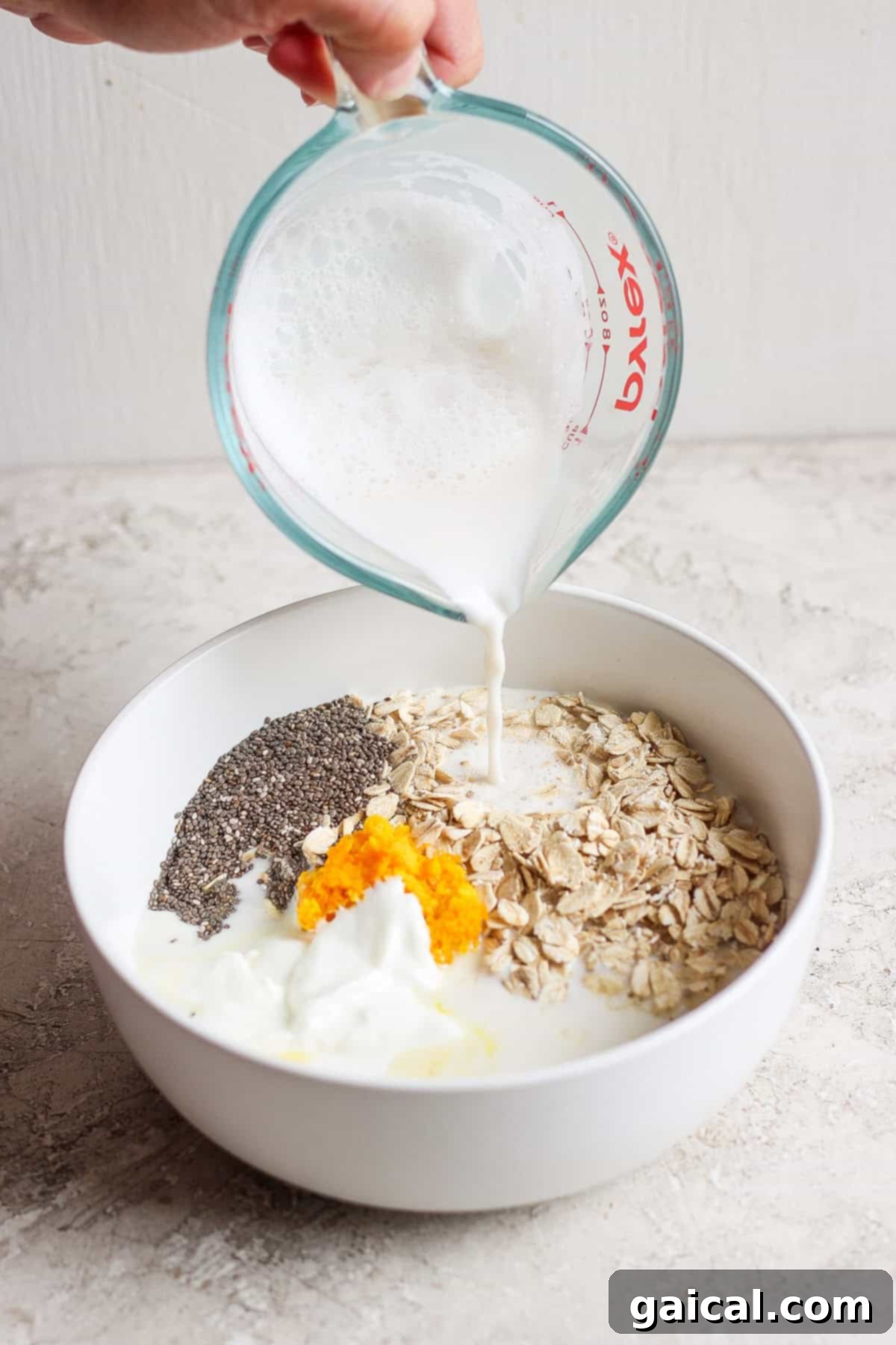 A hand pours milk from a measuring cup into a white bowl containing oats, chia seeds, orange zest, and yogurt, creating the perfect base for creamsicle overnight oats.
