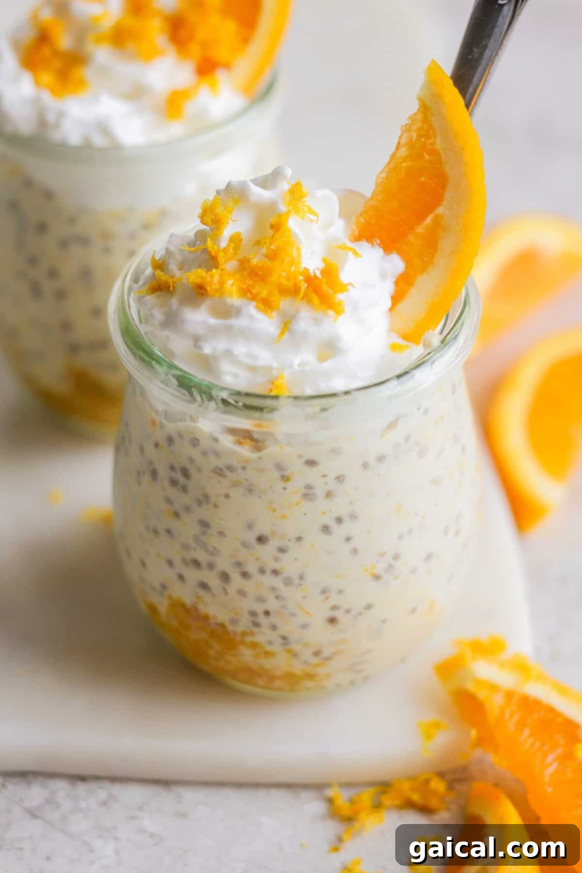 Two small glass jars filled with creamy chia seed pudding, reminiscent of creamsicle overnight oats, are topped with whipped cream, orange zest, and an orange slice. Orange slices on the side complete this delightful treat.