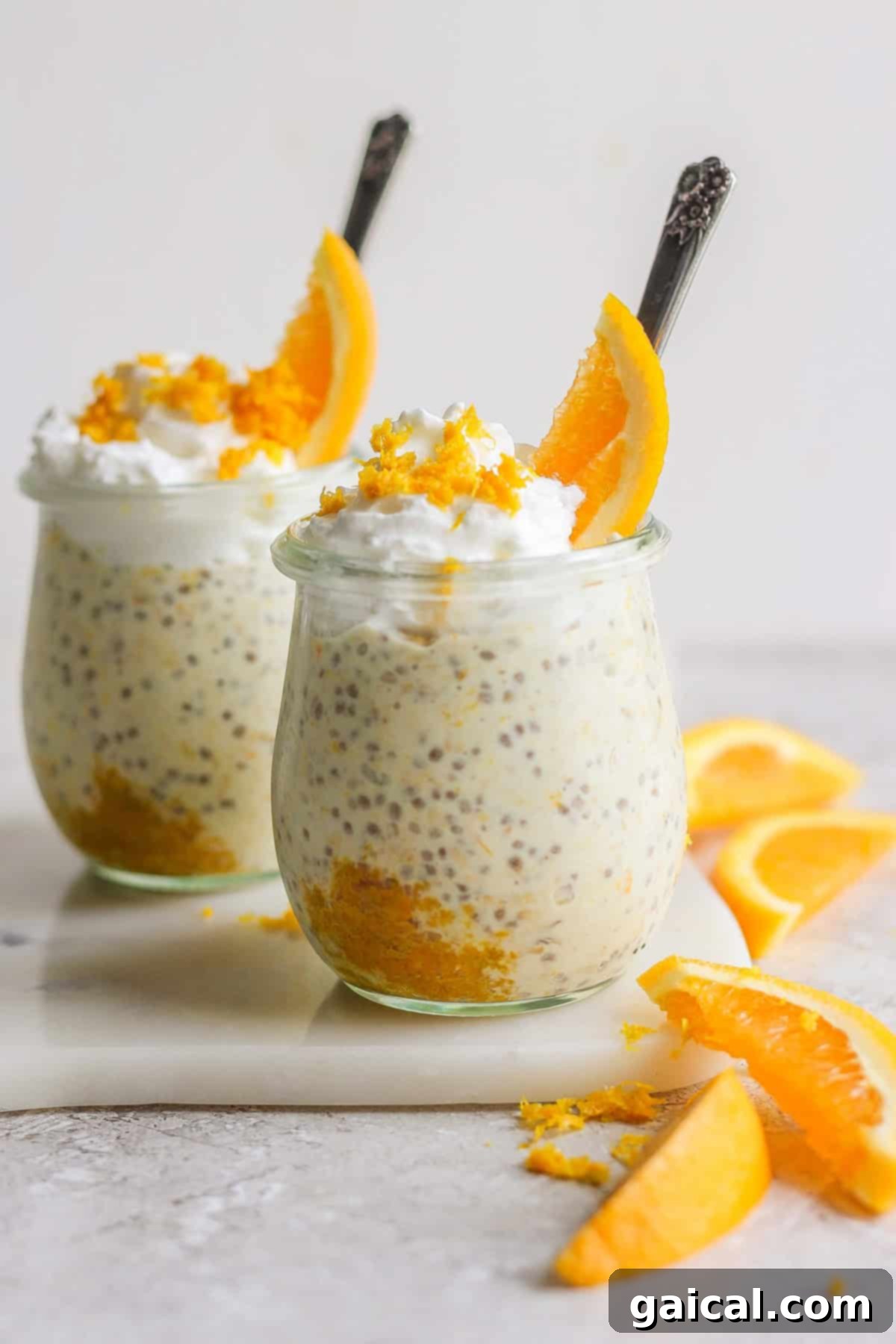Two glass jars filled with creamy creamsicle overnight oats, topped with whipped cream and orange slices, arranged on a white surface with additional orange wedges nearby.