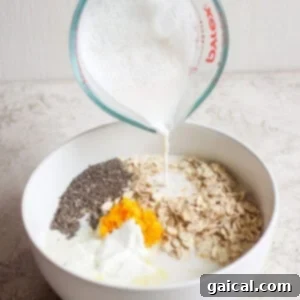 A hand pours milk from a measuring cup into a white bowl containing oats, chia seeds, orange zest, and yogurt, creating the perfect base for creamsicle overnight oats.