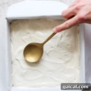 A hand holding a spoon smooths out a white batter for peanut butter banana ice box cake in a parchment-lined square baking pan on a textured countertop.