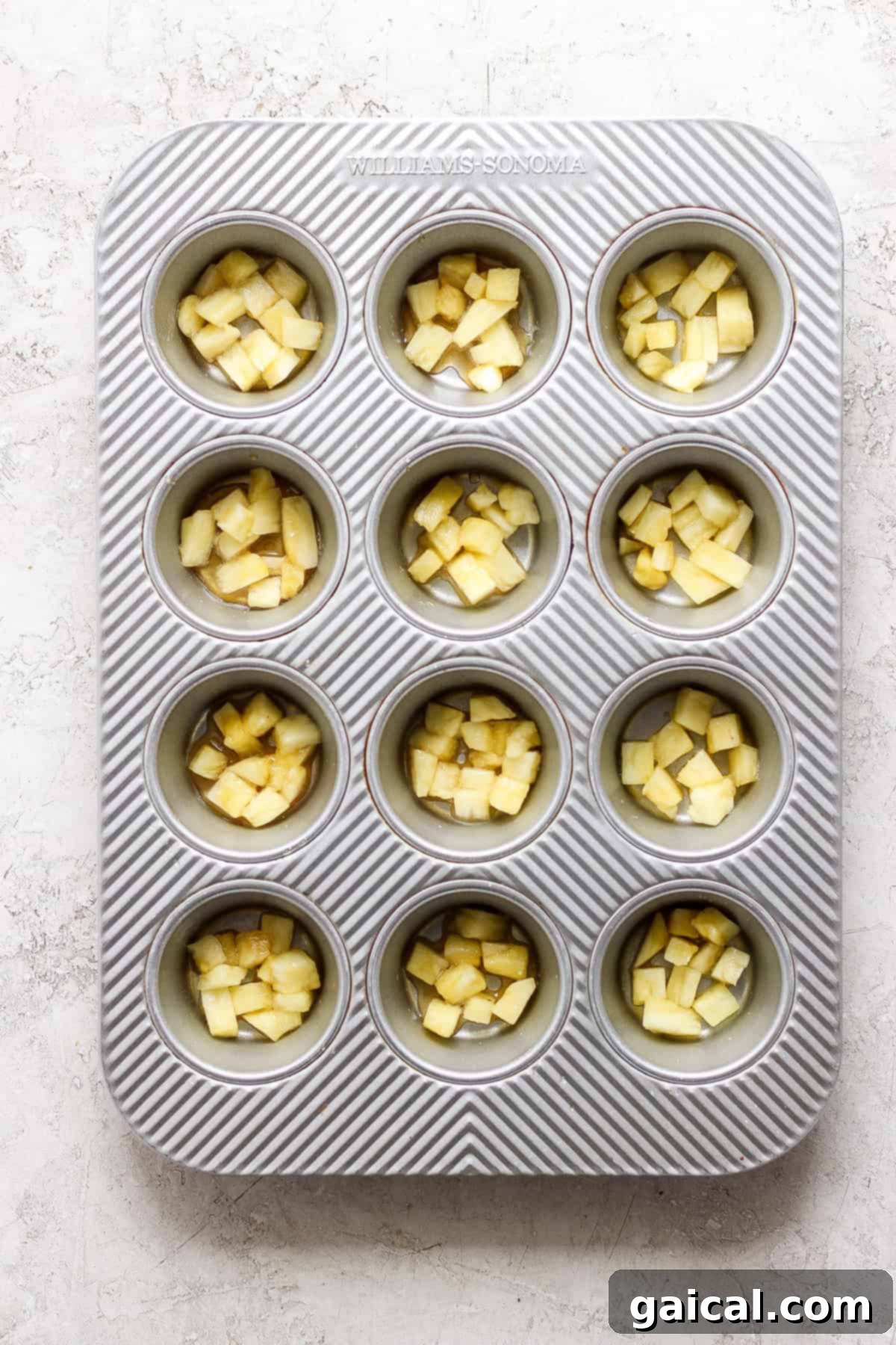 Golden Pineapple Flips 3 A muffin tin containing evenly chopped pineapple chunks in each compartment, set on a light textured surface, ready for batter.
