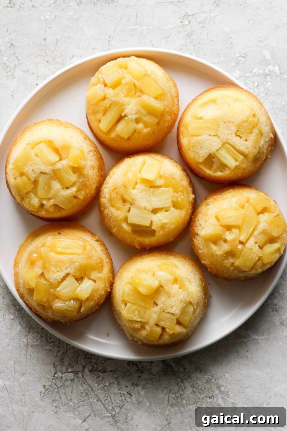 Golden Pineapple Flips 4 A trio of pineapple upside-down mini cakes, each beautifully topped with a pineapple chunk, arranged on a light textured surface.