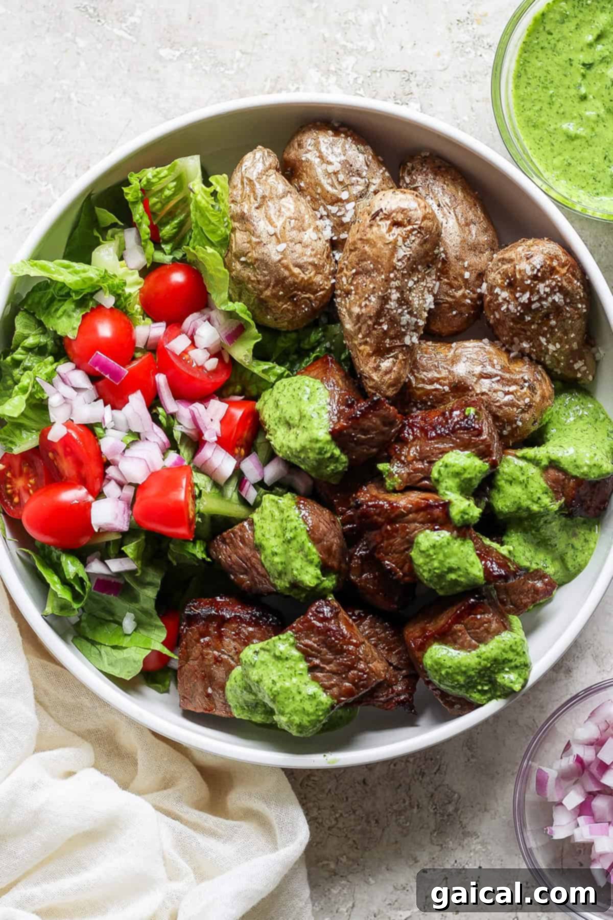 A vibrant bowl featuring crispy grilled steak chunks generously drizzled with a bright green herb sauce, alongside golden roasted potatoes and a fresh side salad of crisp lettuce, juicy cherry tomatoes, and finely chopped red onions.