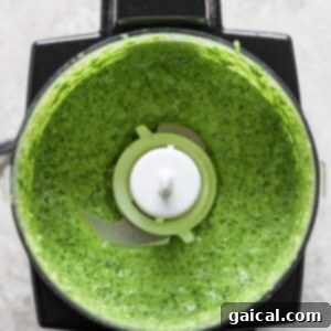 A top-down view of a blender with green sauce inside, featuring visible greens and herbs blended to a smooth consistency.