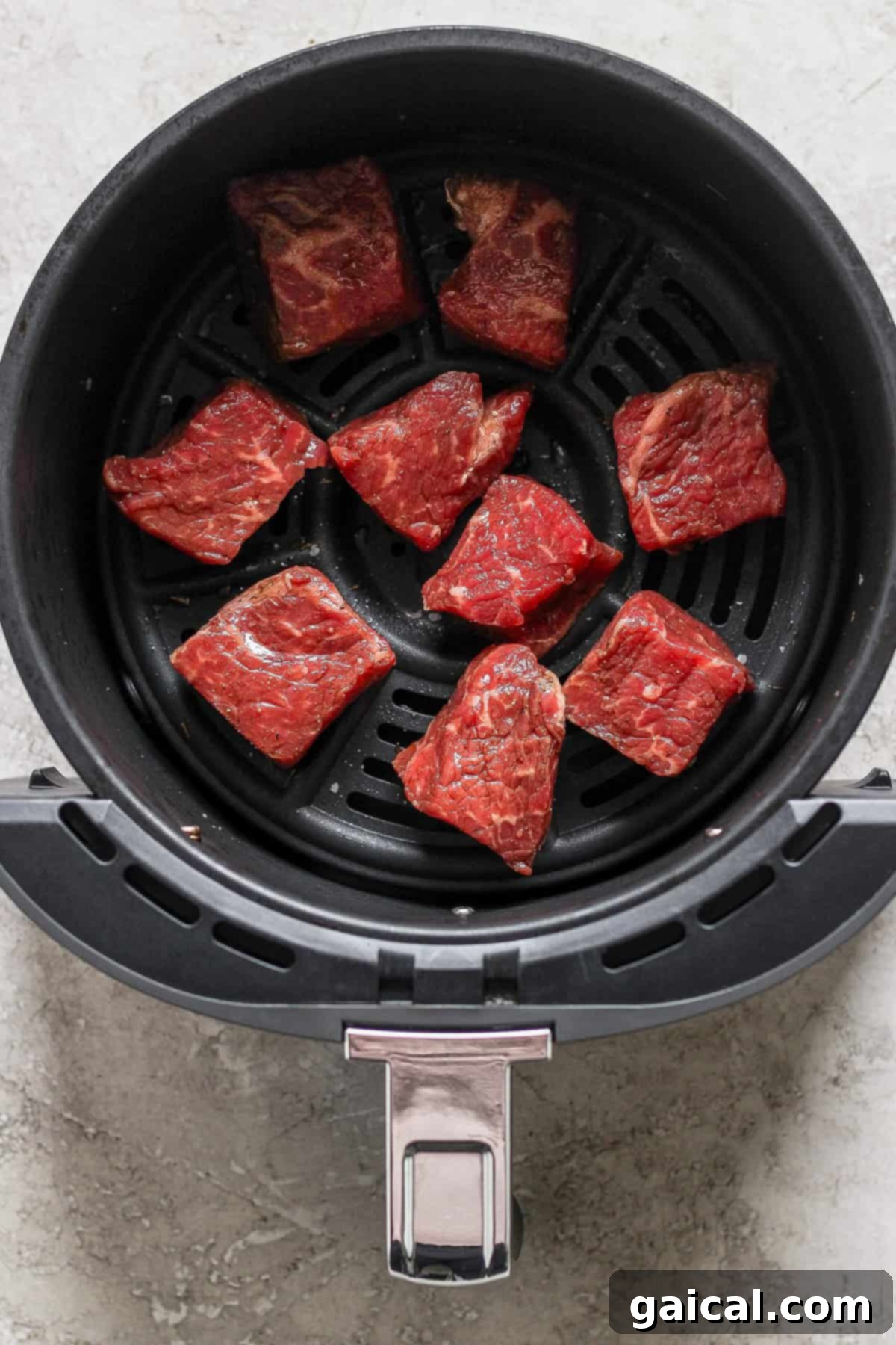 An air fryer basket filled with uniformly cut raw beef pieces, seasoned and ready for cooking, set against a light background.