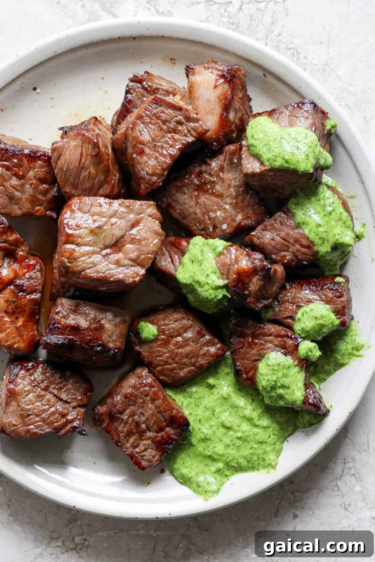 A white plate showcasing succulent grilled steak bites beautifully arranged and topped with a generous dollop of vibrant green herb sauce, ready to be enjoyed.